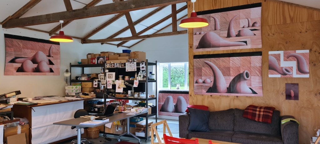 White space with a plywood wall and wooden rafters displaying artwork in pastel peach colours, shelving with cardboard boxes and other containers of various sizes, photographs of artwork, a metal desk and chair with laptop, a gray twoseater couch with folded red tartan blanket over the back of it, and in the foreground a wooden table with red wooden chairs are visible. Two red metal pendant lights light up the studio. 