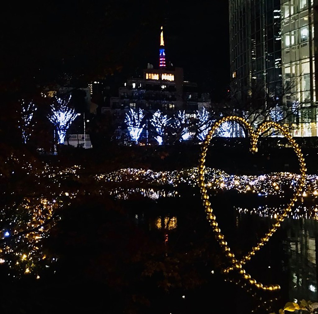 Christmas time in Tokyo at night. A lit up heart shape in the foreground with a row of lit trees and the Tokyo Tower lit up in the background. 